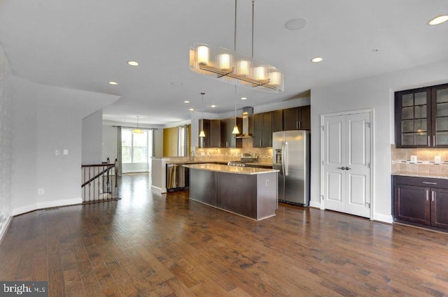 kitchen with dark brown cabinetry, appliances with stainless steel finishes, wall chimney range hood, glass insert cabinets, and pendant lighting