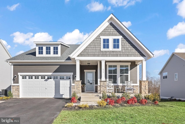 craftsman house featuring a porch, aphalt driveway, a garage, stone siding, and a front lawn