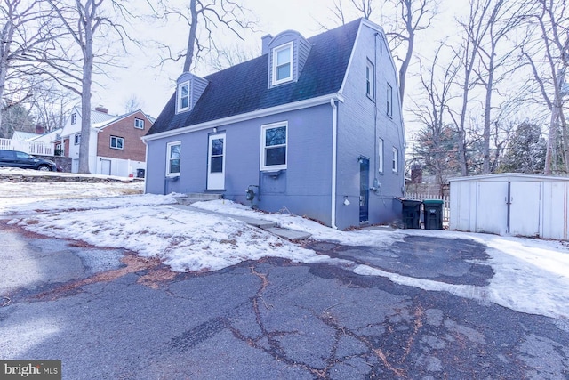 snow covered rear of property with driveway, a gambrel roof, roof with shingles, an outbuilding, and a storage unit