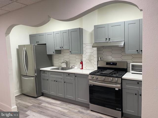 kitchen featuring under cabinet range hood, a sink, light countertops, appliances with stainless steel finishes, and gray cabinets