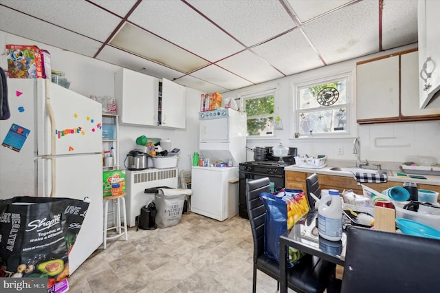 kitchen with a paneled ceiling, freestanding refrigerator, stacked washer / dryer, white cabinets, and a sink