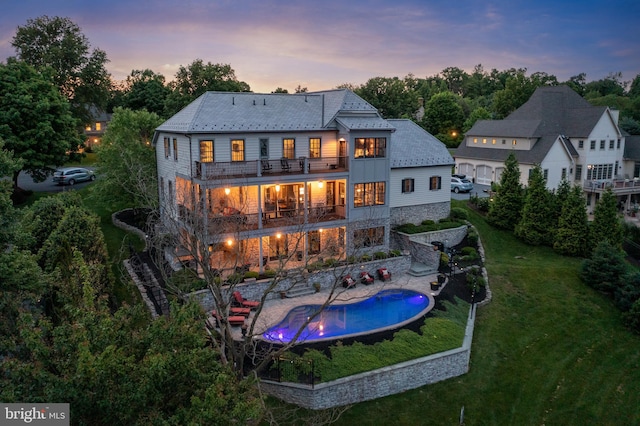 back of property at dusk featuring an outdoor pool, a balcony, a yard, a patio area, and board and batten siding