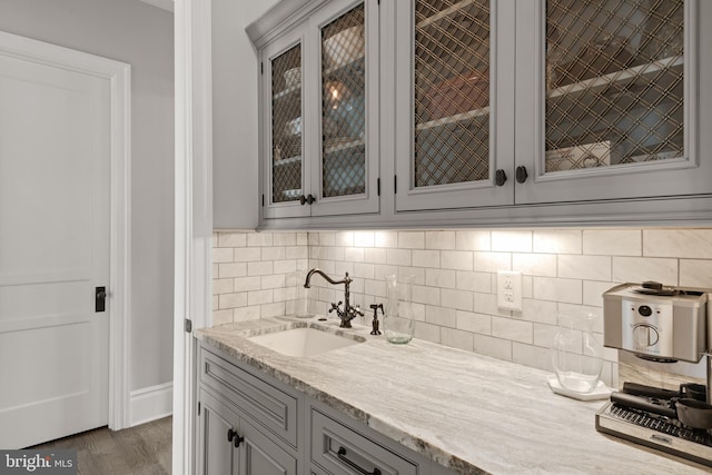 kitchen with decorative backsplash, glass insert cabinets, light stone counters, gray cabinets, and a sink