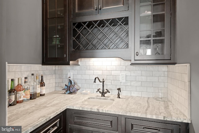 kitchen featuring decorative backsplash, light stone counters, glass insert cabinets, dark brown cabinets, and a sink