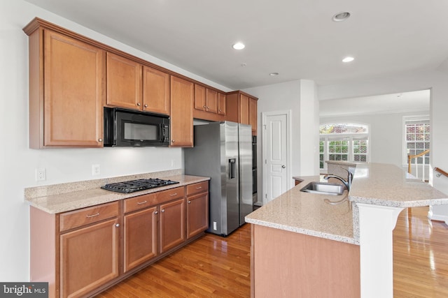 kitchen with brown cabinetry, light stone countertops, a kitchen island with sink, black appliances, and a sink