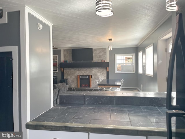 kitchen with a stone fireplace, tile countertops, white cabinets, and crown molding