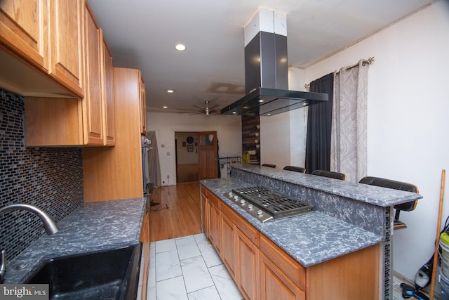 kitchen featuring island range hood, stainless steel gas cooktop, a sink, decorative backsplash, and brown cabinetry