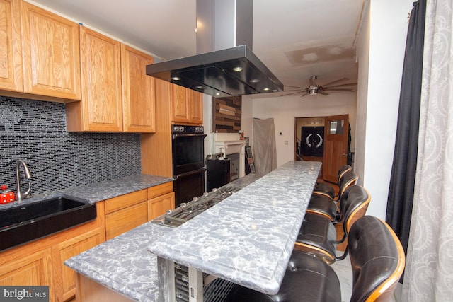 kitchen with dobule oven black, a ceiling fan, decorative backsplash, light stone countertops, and island exhaust hood