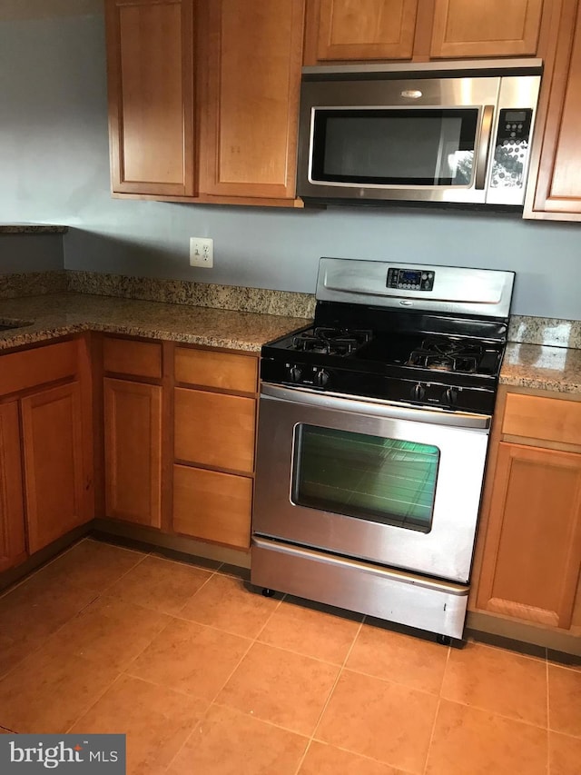 kitchen with light tile patterned floors, appliances with stainless steel finishes, dark stone counters, and brown cabinets