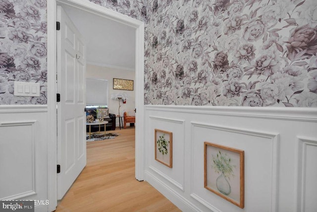 hallway with a wainscoted wall, ornamental molding, light wood-style flooring, and wallpapered walls