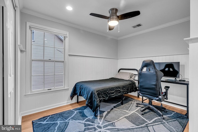 bedroom featuring ceiling fan, a wainscoted wall, wood finished floors, visible vents, and crown molding