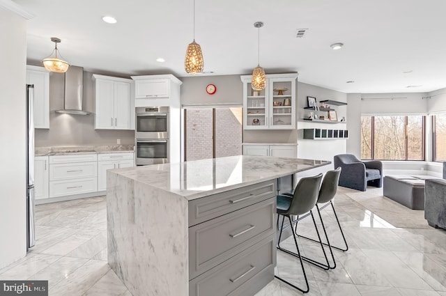 kitchen featuring white cabinets, wall chimney exhaust hood, appliances with stainless steel finishes, a center island, and light stone countertops