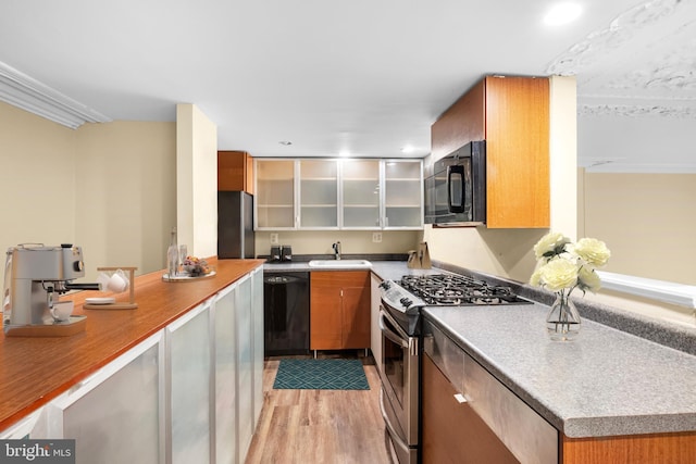 kitchen featuring light wood finished floors, glass insert cabinets, brown cabinetry, a sink, and black appliances