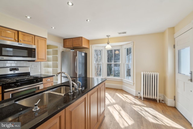 kitchen featuring stainless steel appliances, light wood-type flooring, backsplash, radiator, and dark stone countertops