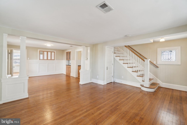 unfurnished living room featuring light wood finished floors, visible vents, stairs, a healthy amount of sunlight, and ornate columns