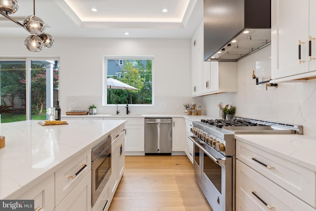 kitchen featuring light stone counters, a tray ceiling, appliances with stainless steel finishes, white cabinetry, and wall chimney range hood