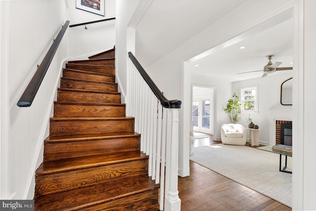 stairway featuring a ceiling fan, a brick fireplace, baseboards, and wood finished floors