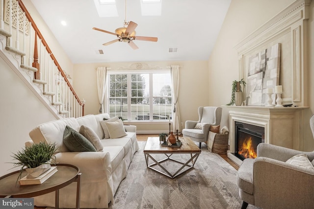 living room featuring ceiling fan, visible vents, stairs, lofted ceiling with skylight, and a glass covered fireplace