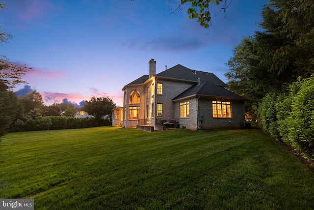 back of house at dusk featuring brick siding, a chimney, and a yard