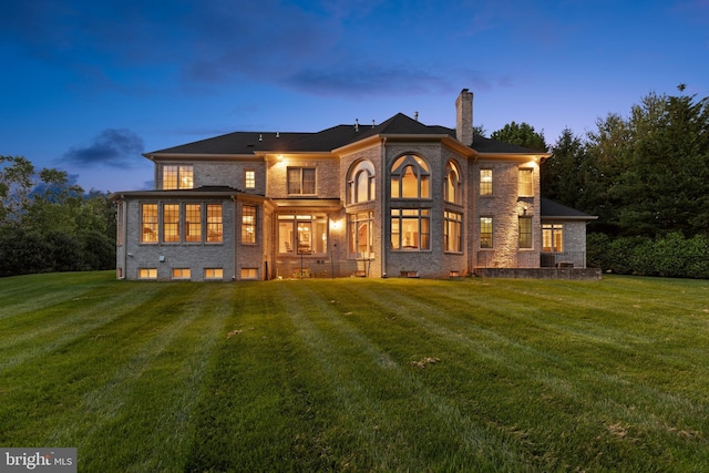 back of property at dusk featuring brick siding, a lawn, and a chimney