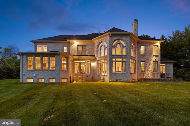 back of property at dusk with brick siding, a yard, and a chimney