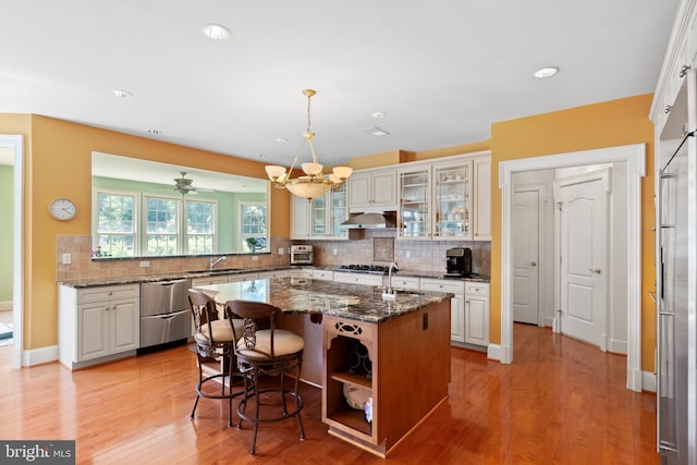 kitchen featuring a center island with sink, decorative backsplash, glass insert cabinets, a sink, and exhaust hood