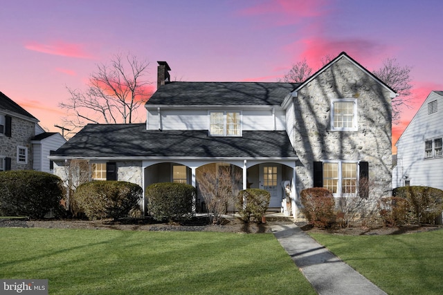 view of front of home with covered porch, a shingled roof, a chimney, and a front lawn