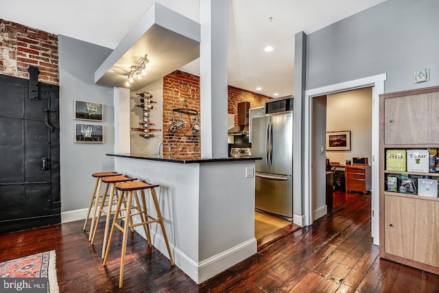 kitchen with a peninsula, dark wood-style flooring, wall chimney range hood, freestanding refrigerator, and dark countertops