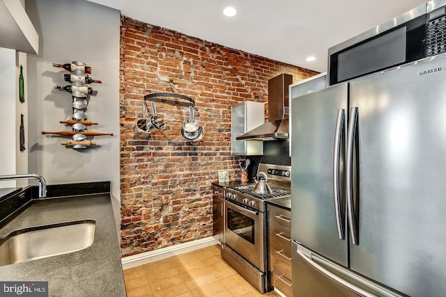 kitchen with dark countertops, brick wall, appliances with stainless steel finishes, wall chimney range hood, and a sink