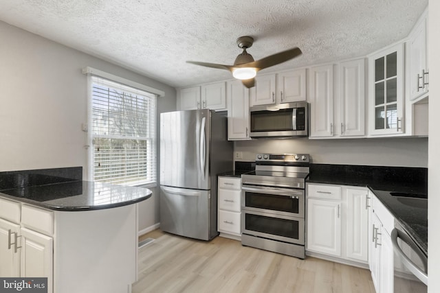 kitchen with dark countertops, appliances with stainless steel finishes, glass insert cabinets, white cabinetry, and light wood-type flooring