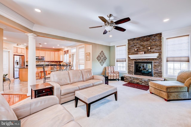 living area with ceiling fan, a stone fireplace, light carpet, ornamental molding, and ornate columns