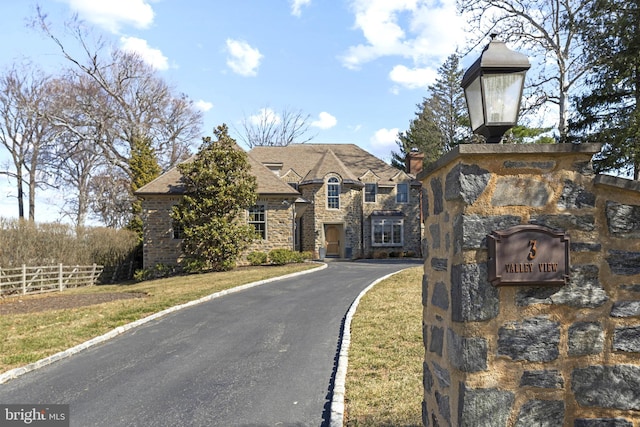 view of front facade featuring aphalt driveway, stone siding, a chimney, and fence