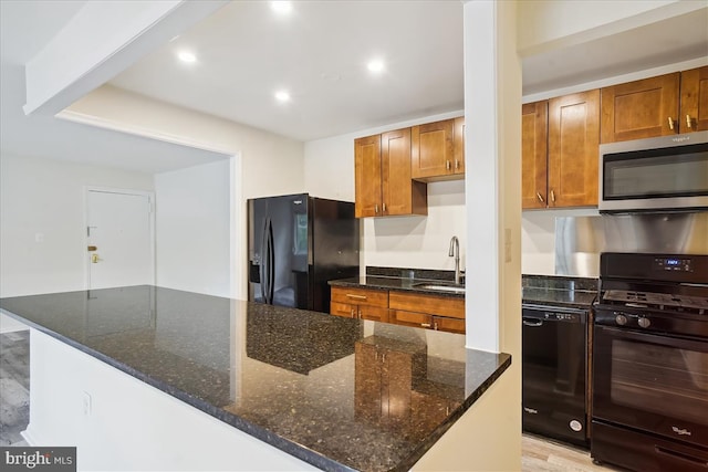 kitchen with brown cabinets, recessed lighting, a sink, dark stone countertops, and black appliances