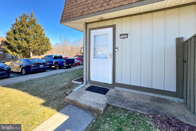 entrance to property featuring a shingled roof, mansard roof, a yard, uncovered parking, and board and batten siding