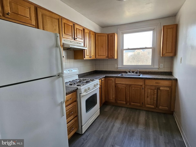 kitchen featuring dark countertops, brown cabinetry, a sink, white appliances, and under cabinet range hood