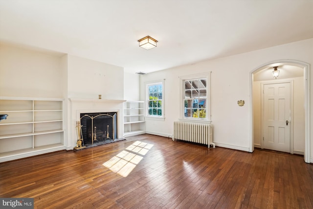 unfurnished living room with arched walkways, a fireplace with raised hearth, baseboards, hardwood / wood-style floors, and radiator heating unit