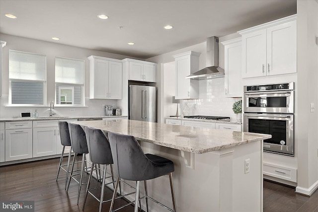 kitchen featuring a center island, appliances with stainless steel finishes, white cabinetry, a sink, and wall chimney exhaust hood