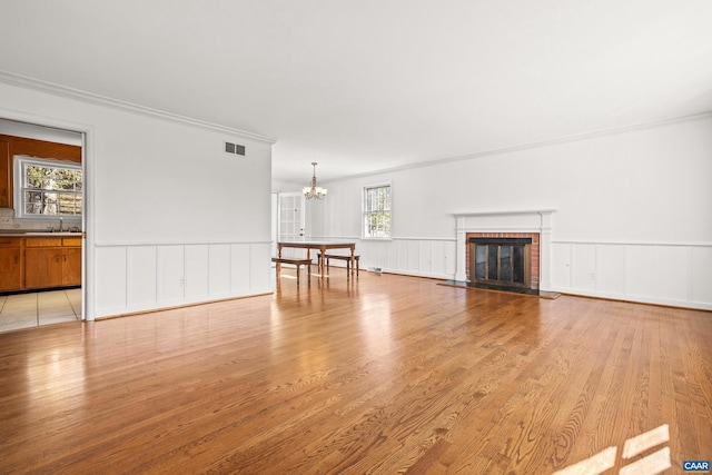 unfurnished living room featuring a chandelier, light wood-style flooring, visible vents, ornamental molding, and a brick fireplace