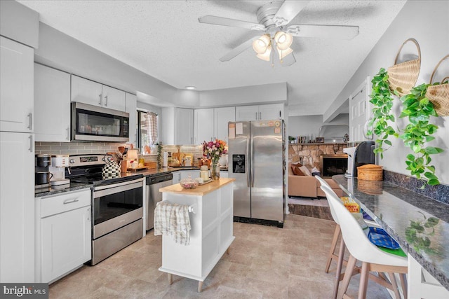 kitchen with decorative backsplash, appliances with stainless steel finishes, white cabinetry, a stone fireplace, and a textured ceiling