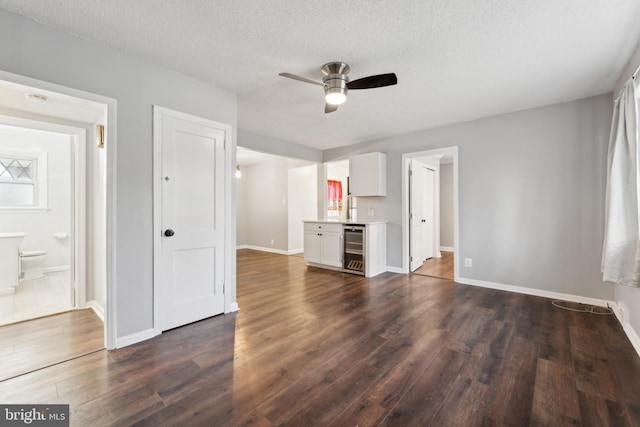 unfurnished living room featuring a textured ceiling, beverage cooler, dark wood-type flooring, a ceiling fan, and baseboards