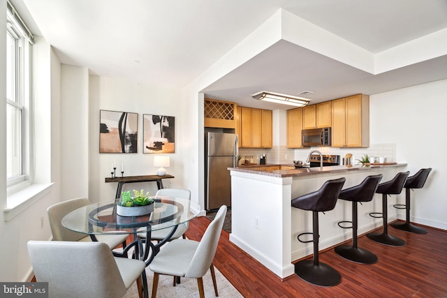 kitchen with dark wood-style flooring, a peninsula, stainless steel appliances, light brown cabinetry, and backsplash