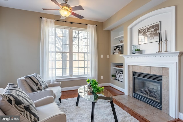 living area featuring built in shelves, baseboards, a tiled fireplace, and a ceiling fan