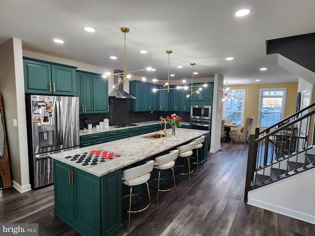 kitchen with a kitchen island with sink, stainless steel appliances, a sink, hanging light fixtures, and wall chimney exhaust hood