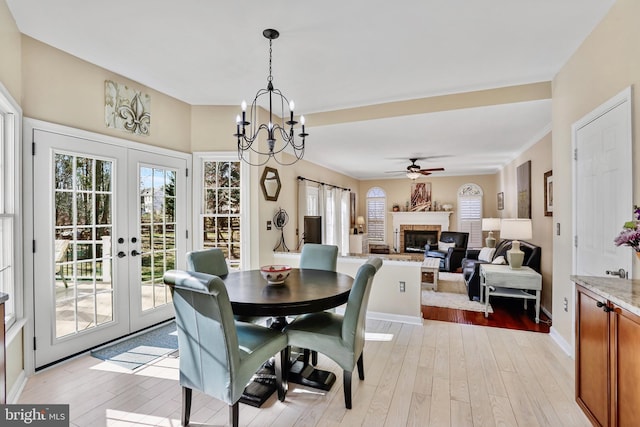 dining room with ceiling fan with notable chandelier, baseboards, light wood-style floors, french doors, and a brick fireplace