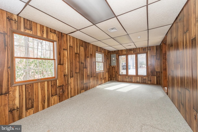 carpeted empty room featuring electric panel, visible vents, wooden walls, and a drop ceiling