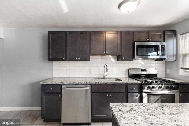 kitchen featuring dark brown cabinetry, a sink, light stone countertops, stainless steel appliances, and backsplash