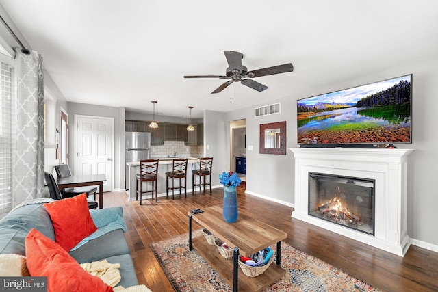 living area featuring dark wood-style flooring, visible vents, a glass covered fireplace, ceiling fan, and baseboards