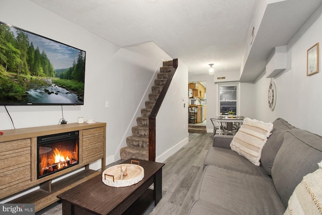 living room with a textured ceiling, wood finished floors, baseboards, stairs, and a lit fireplace