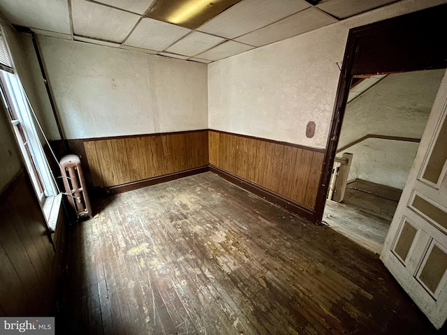 empty room featuring wooden walls, wainscoting, dark wood-style floors, radiator heating unit, and a paneled ceiling