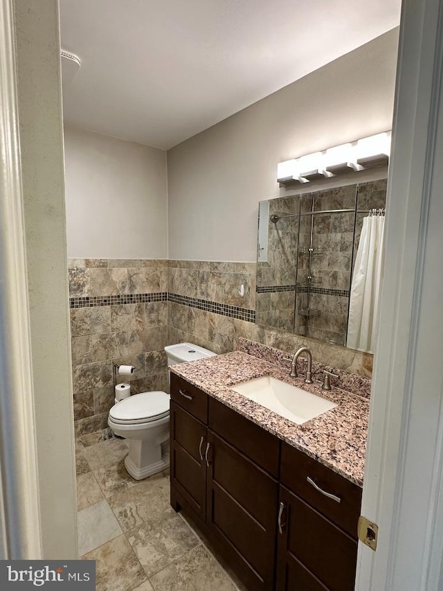 bathroom featuring a wainscoted wall, tile walls, toilet, vanity, and tiled shower